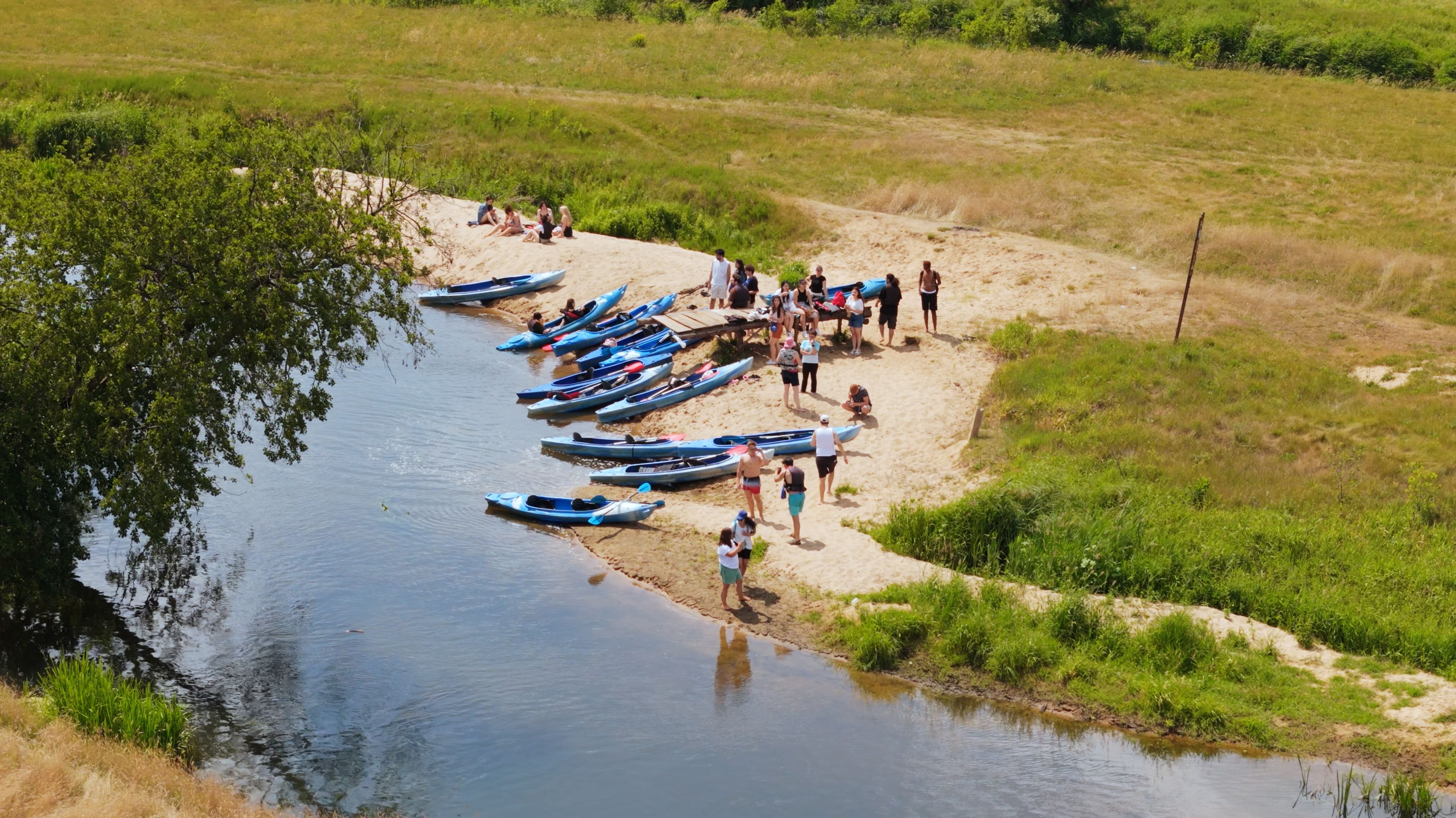 Grupa studentów przygotowuje kajaki na brzegu rzeki przed spływem.